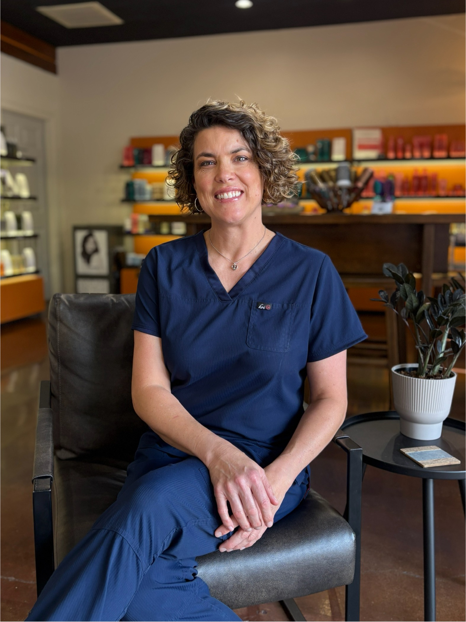 A woman with short curly hair, wearing navy blue scrubs, sits on a chair in a modern, well-lit room with shelves and plants in the background.