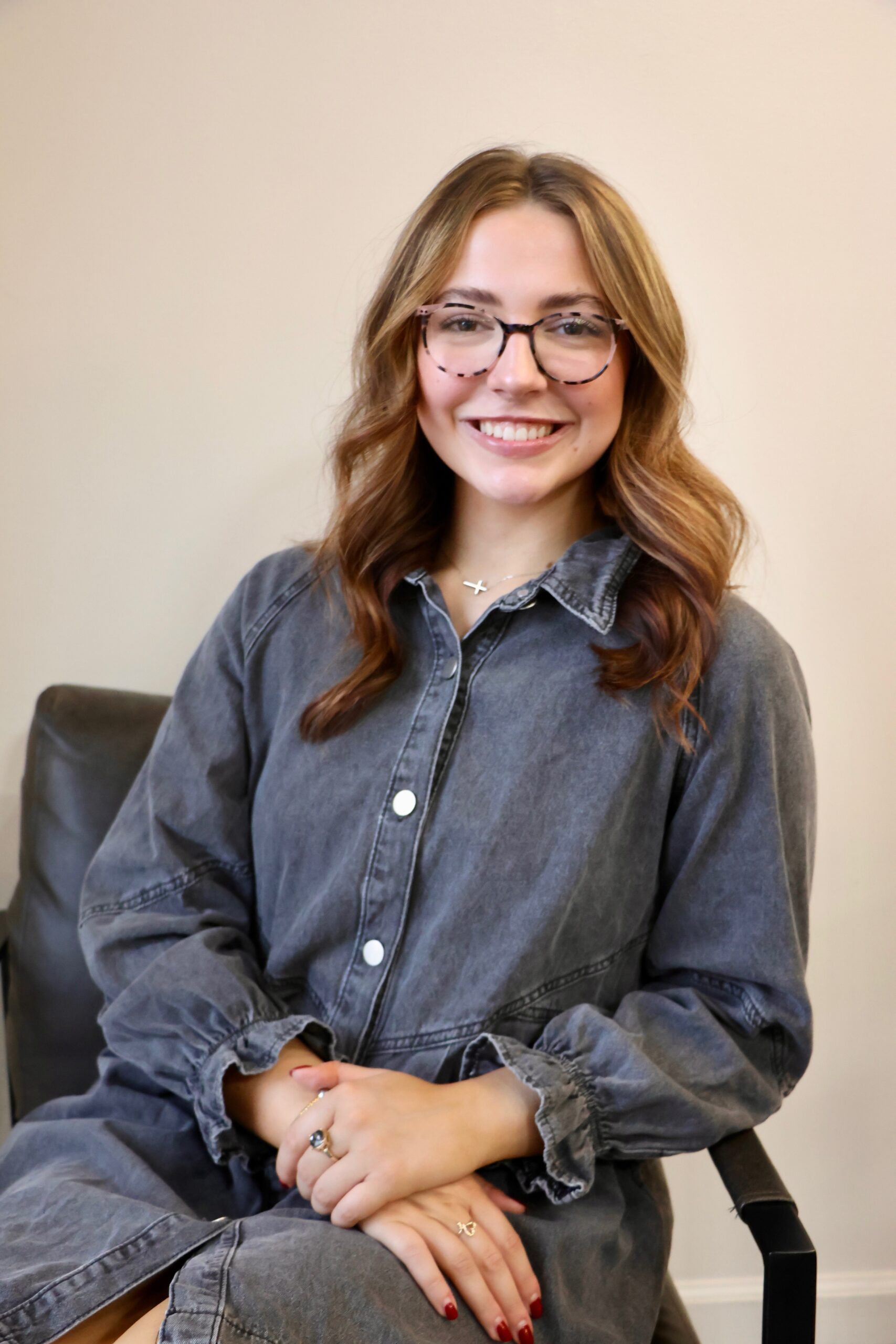 A person with long hair and glasses, wearing a gray button-up dress, smiles while seated in an armchair against a plain background.