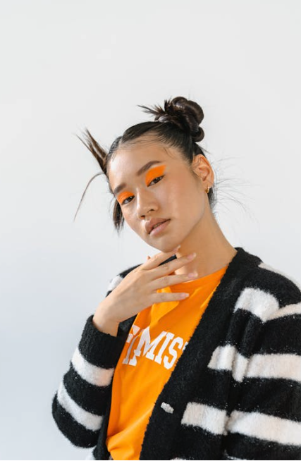 A person with a double bun hairstyle poses confidently. They wear a black and white striped sweater over an orange shirt. Their bright orange eyeshadow matches the shirt.