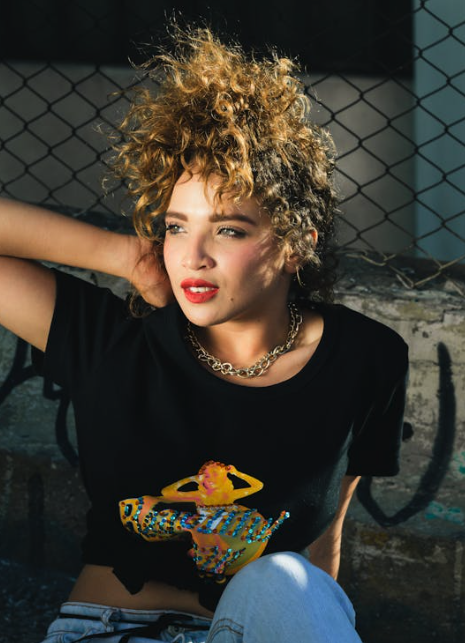 A person with curly hair leans against a graffiti-covered wall, wearing a black t-shirt with a colorful graphic and a chain necklace.