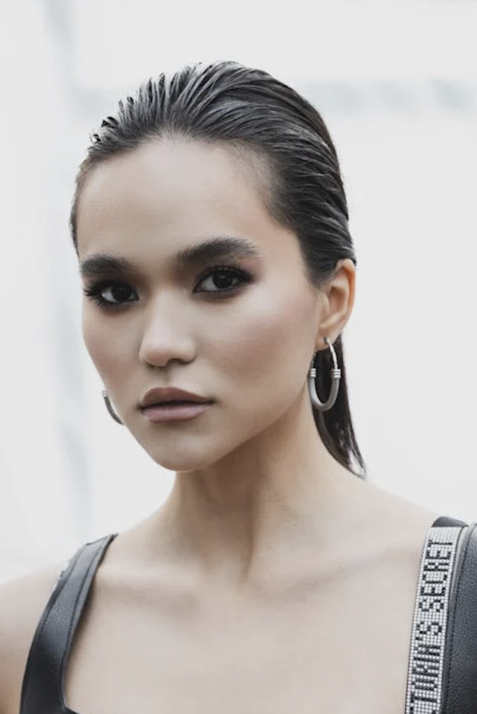 Person with slicked-back hair wearing hoop earrings and a black outfit, looking directly ahead.