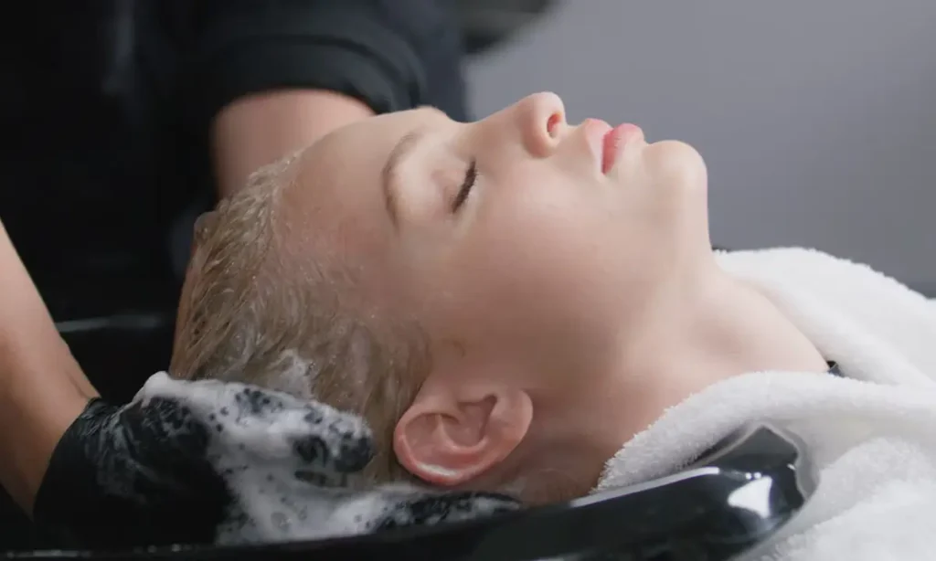 A person with closed eyes is having their hair washed at a salon, with shampoo lather visible and a towel around their neck.