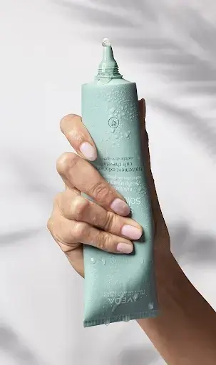 A hand holding a light green tube of cream with a small drop of product at the opening, against a white background—perfect for enhancing your self-care and wellness rituals.