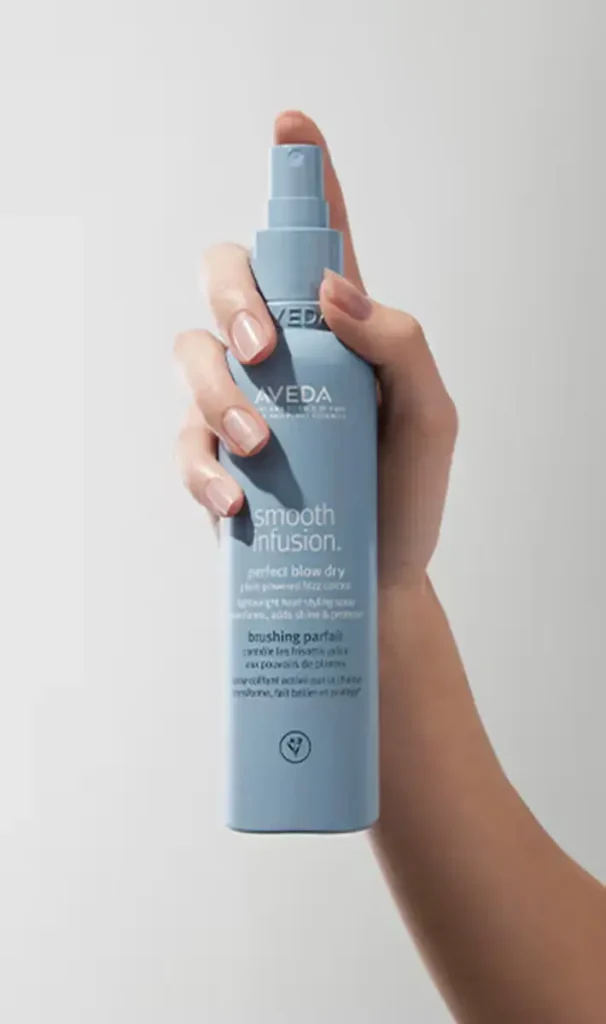 A hand holding a blue bottle of Aveda Smooth Infusion Perfect Blow Dry spray against a plain light background, perfect for elevating your self-care and wellness rituals.