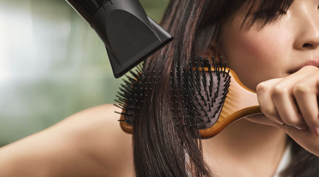 A person blow-dries their straight, shoulder-length hair while brushing it with a wooden paddle brush, following essential winter hair care for smooth and healthy-looking strands.