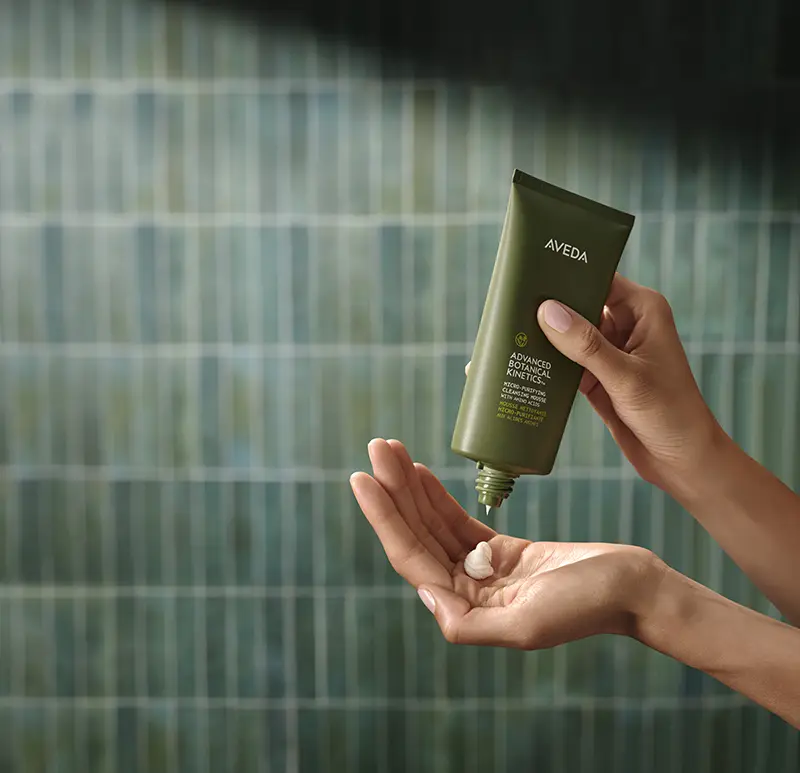 A person squeezes a small amount of Aveda product from a green tube into their hand against a tiled background.