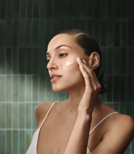 A woman applies a cream to her cheek with her fingers while standing in front of a green tiled wall, starting her spring hair and skin prep routine.