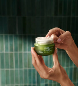 A person holds an open jar of Aveda Advanced Botanical Repair Intensive Strengthening Masque against a green tiled background, perfect for spring hair and skin prep with trusted Aveda hair care.