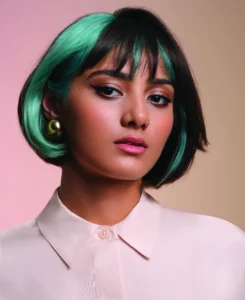 A woman with short black and green hair, wearing a light blouse, gold earrings, and a nose ring, poses against a pastel background.