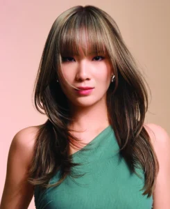 Woman with straight, layered brown hair and bangs, wearing a green sleeveless top, poses against a neutral background.