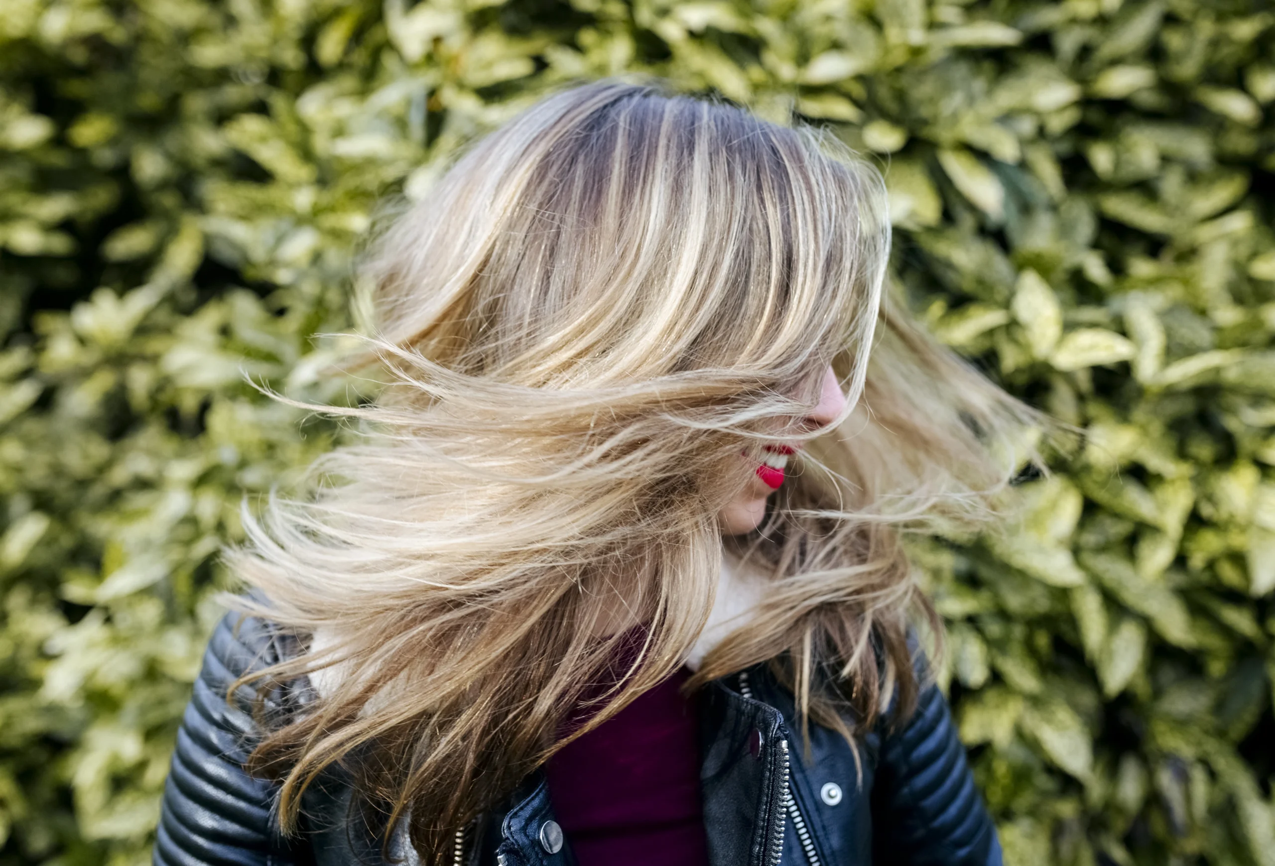 A person with long blonde hair wearing a black jacket turns their head, causing their hair to fan out, with green leaves in the background.