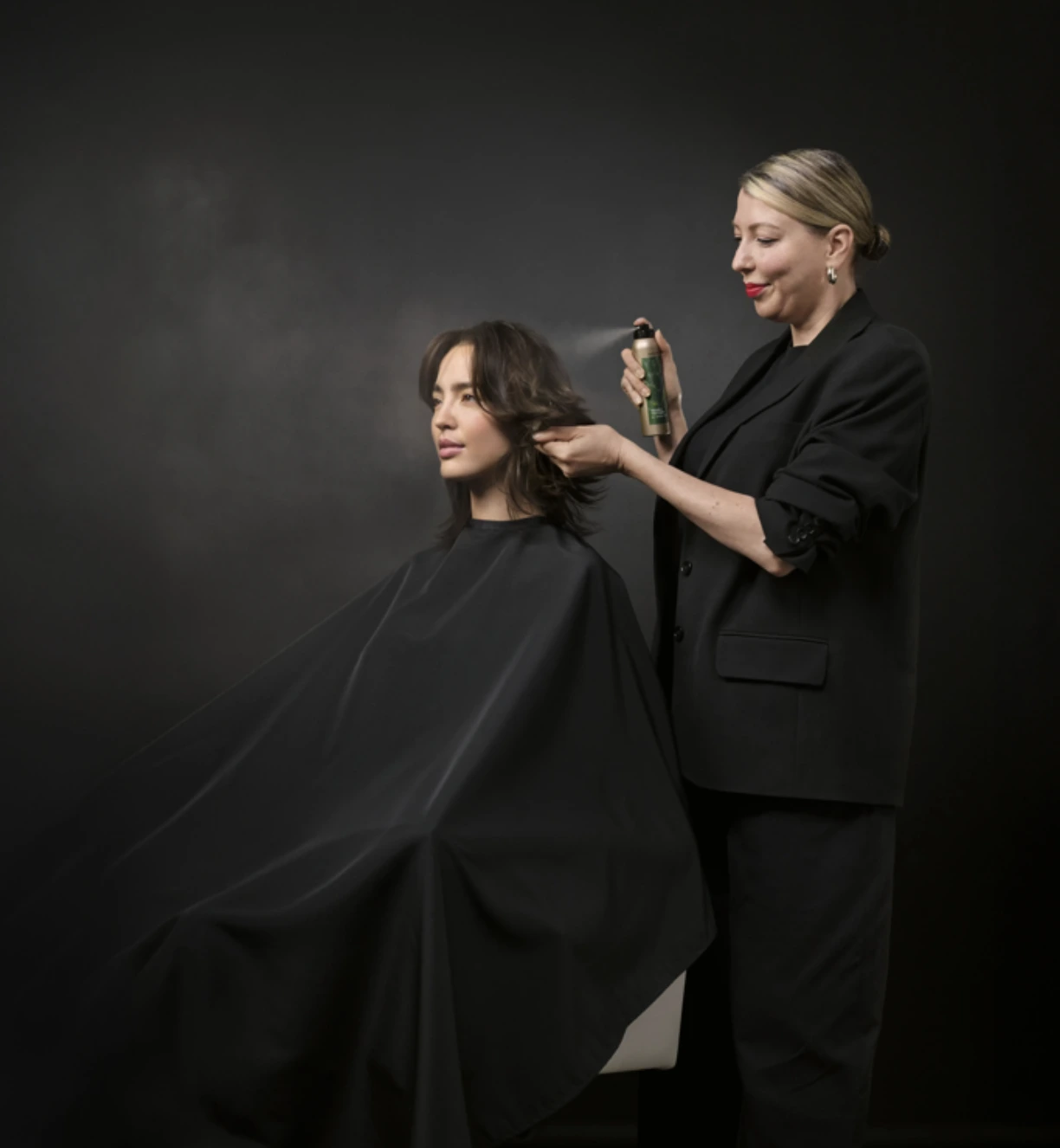 A hairstylist dressed in black applies hairspray to a client's hair, who is seated and covered with a black cape, against a dark background.