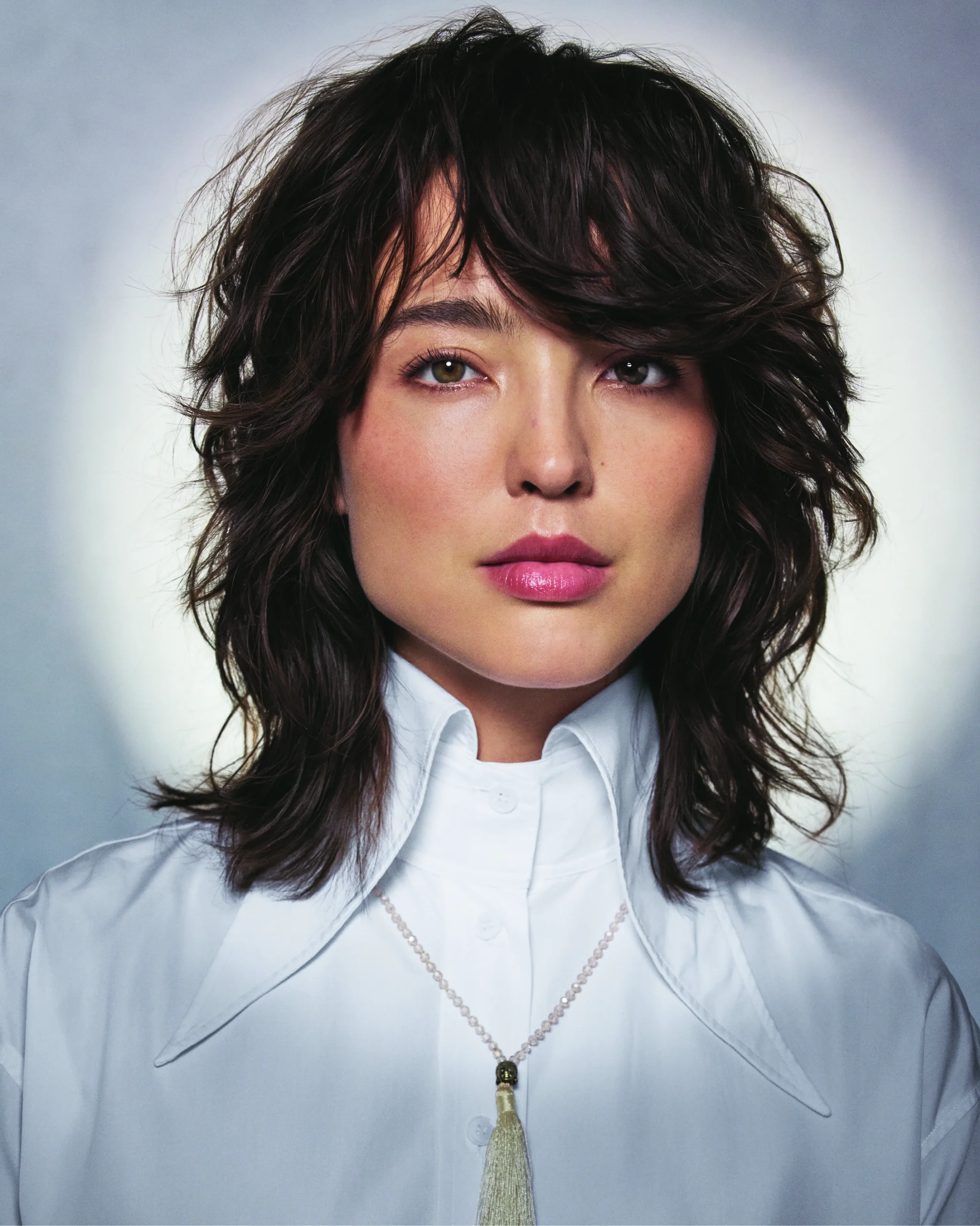 Person with tousled, layered dark hair wearing a white collared shirt and a necklace, posed against a neutral background with a soft halo effect.