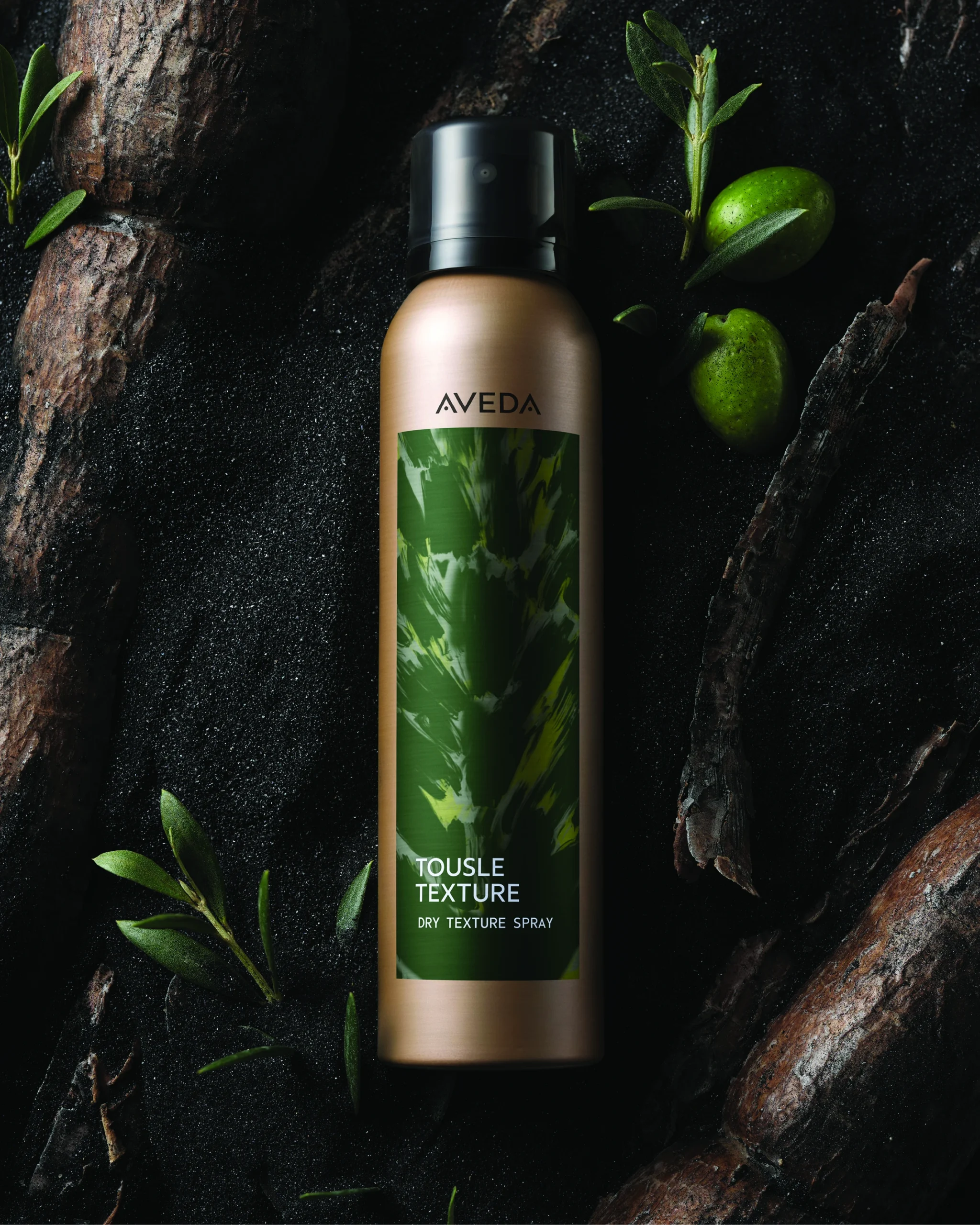 A can of Aveda Tousle Texture Dry Texture Spray is placed on a dark surface with green leaves, olives, and branches scattered around it.